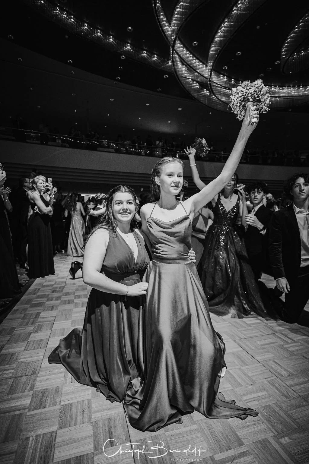 Zwei junge Frauen in eleganten Abendkleidern beim Tanzen auf einer festlichen Veranstaltung, eine mit hochgelegtem Arm, die andere lächelnd neben ihr, im Hintergrund weitere Tänzer und ein dunkler Veranstaltungsraum.