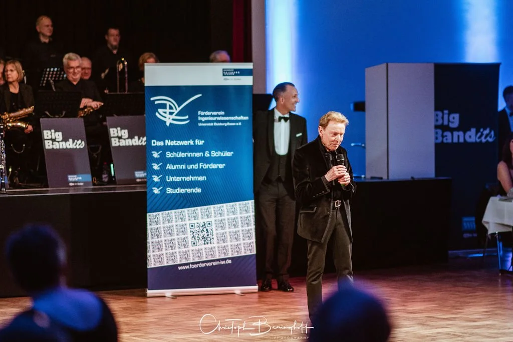 Ein Mann hält eine Handmikrofon bei einer Veranstaltung mit einer Band im Hintergrund, die auf der Bühne sitzt, und einem Banner mit Informationen über den Förderverein Ingenieurwissenschaften.