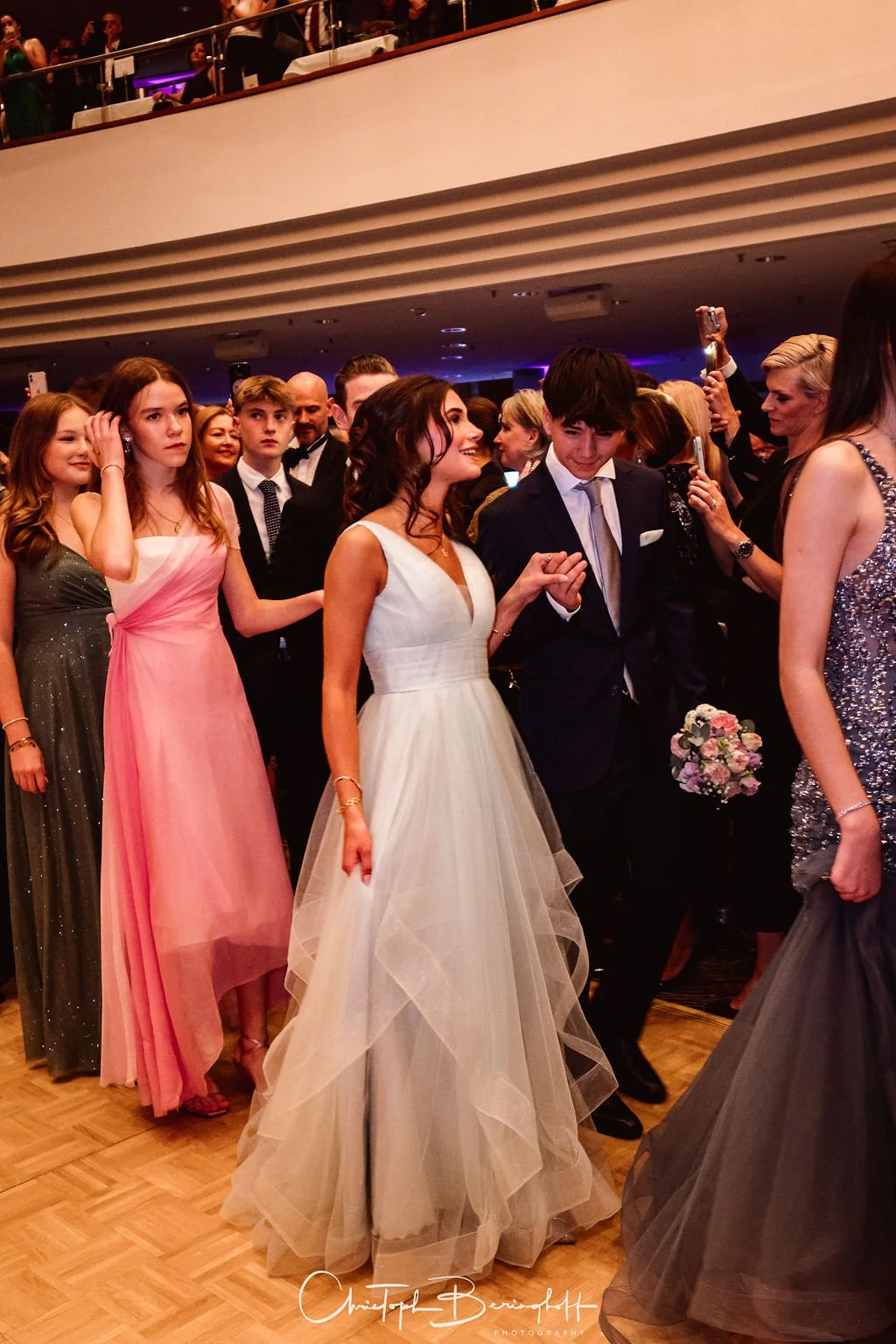 Junge Leute in eleganten Kleidern und Anzügen bei einer Feier, vermutlich ein Abschlussball, tanzen und lachen in einem festlichen Raum.
