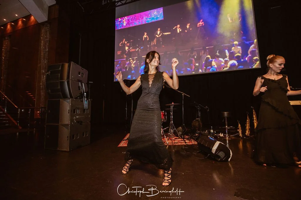 Zwei Frauen tanzen auf einer Bühne vor großer Videoleinwand mit einem Publikum im Hintergrund, während eines Live-Auftritts mit Musik. Die Bühne ist mit Musikinstrumenten ausgestattet.