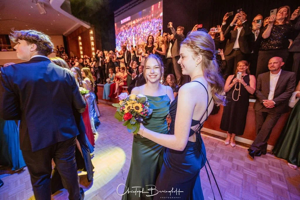 Zwei Frauen in eleganten Kleidern des Kulturpreises bei einer Feier, umgeben von Gästen und Fotografen.