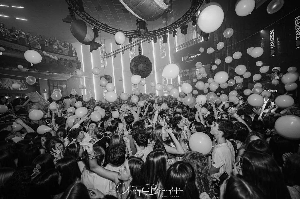 People at a party in a dark room, surrounded by balls and balloons, with beams of light and a dance floor in the background.
