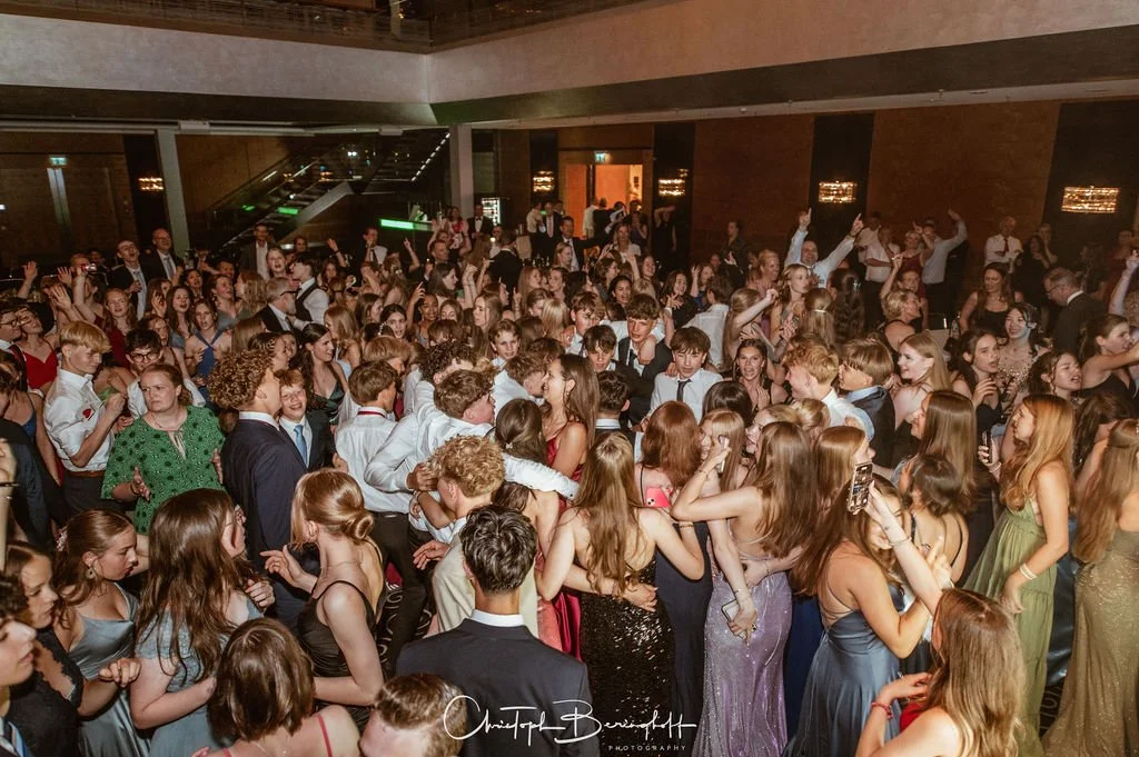 Menschen tanzen und feiern auf einer Party in einem großen Saal, überwiegend junge Erwachsene in festlicher Kleidung.