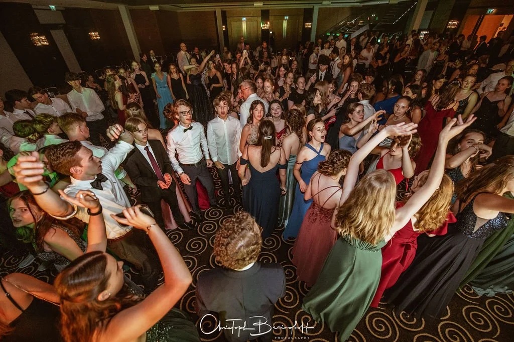 Eine Gruppe junger Menschen, die bei einer Ballnacht tanzen und feiern, viele tragen elegante Kleider und Anzüge, im Hintergrund eine große Menschenmenge in einem festlich dekorierten Saal.