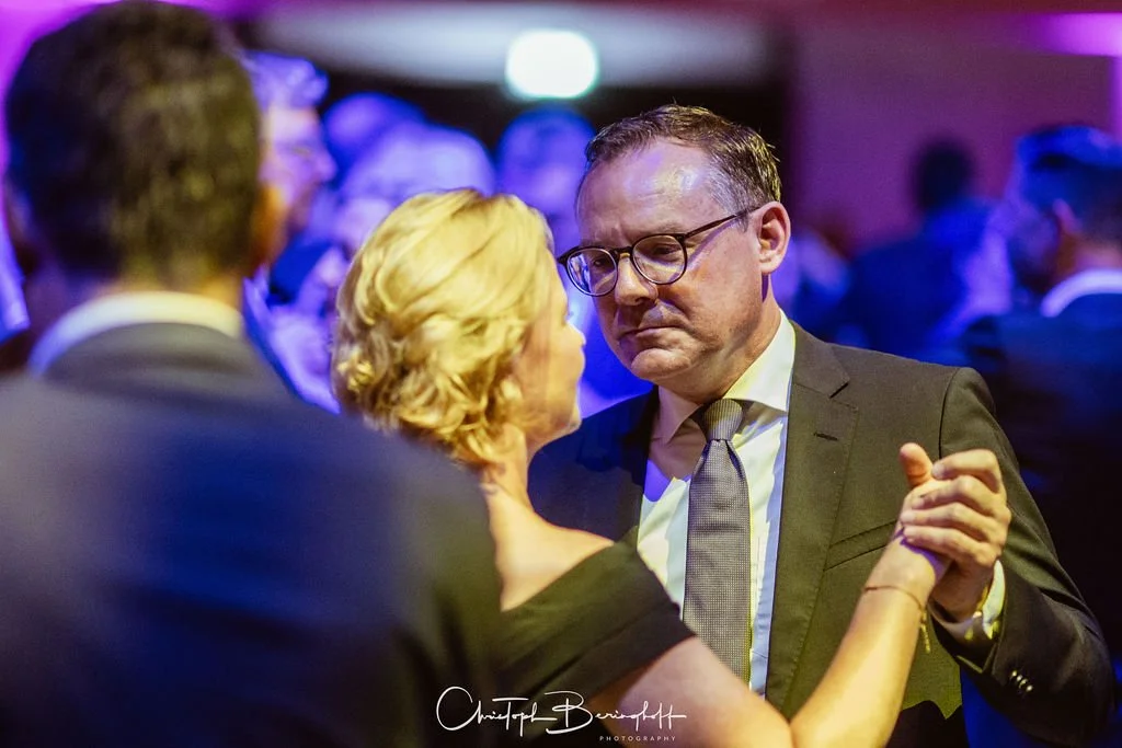 Ein Mann im Anzug und Brille tanzt mit einer älteren Dame auf einer Party mit dunklem, buntem Licht im Hintergrund.