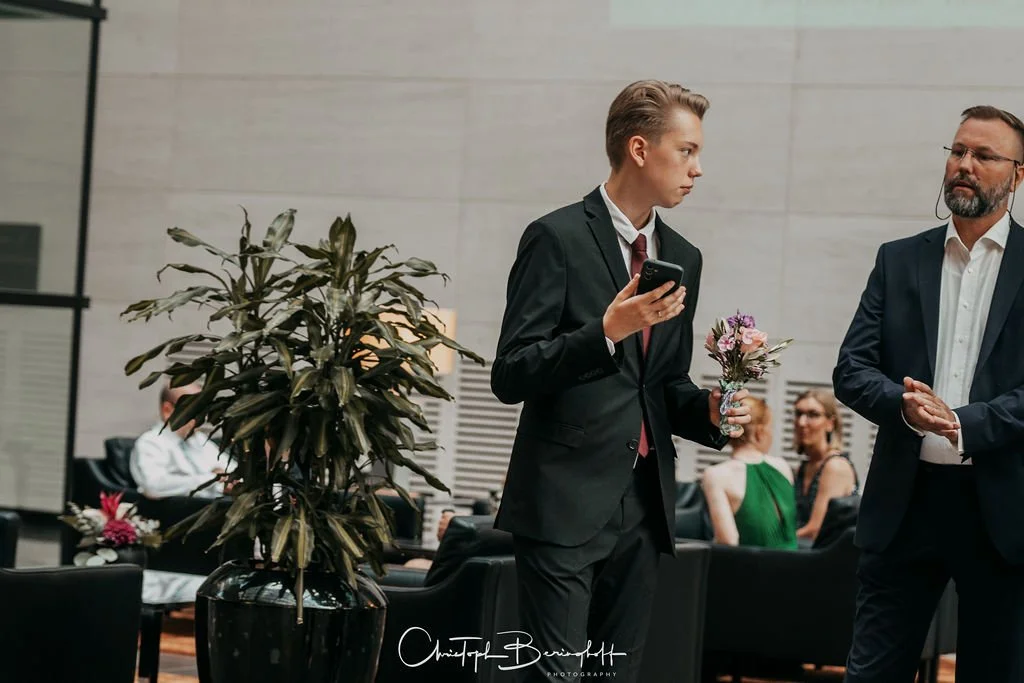 Zwei Männer in Businesskleidung unterhalten sich in einem Lounge-Bereich, einer hält einen Blumenstrauß und ein Smartphone, im Hintergrund sind weitere Personen und Pflanzen.