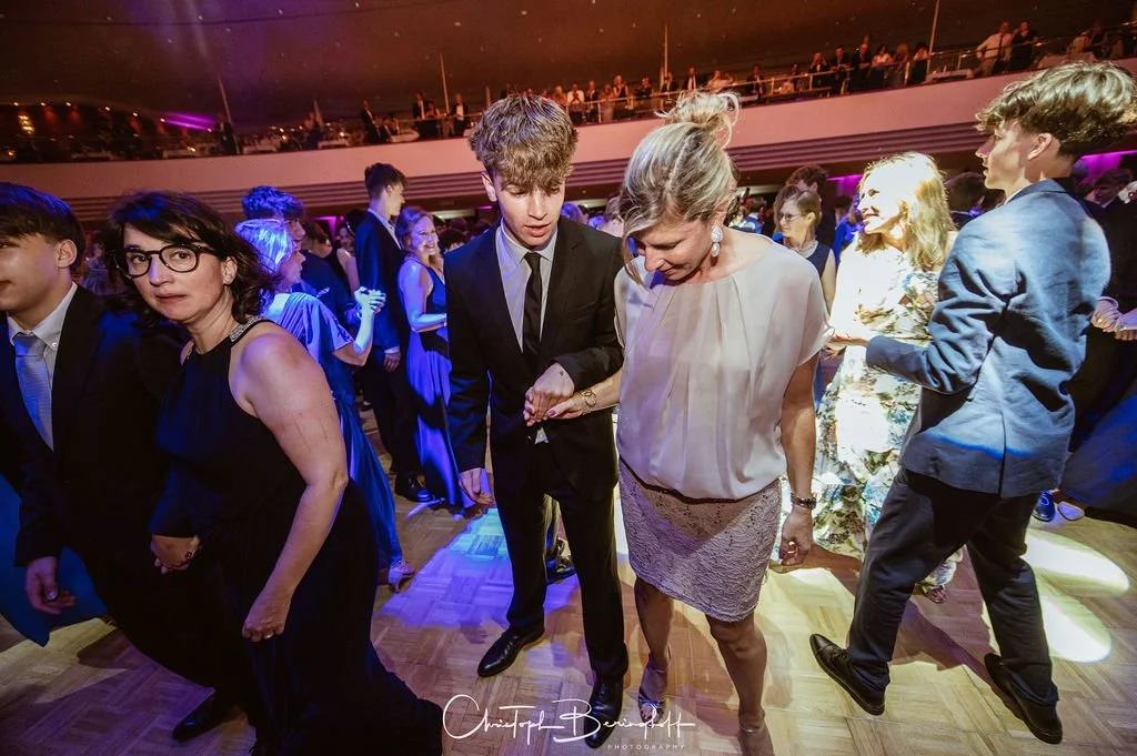 Menschen tanzen auf einer festlichen Veranstaltung, viele im formellen Kleid, im Hintergrund eine Zuschauertribüne mit weiteren Gästen, bunte Beleuchtung am Abend.