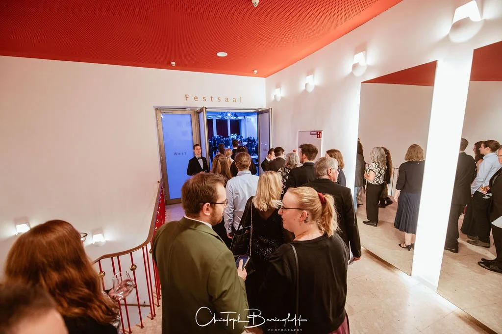 Menschen warten in einer eleganten Halle mit weißer Wand und rotem Decke vor dem Eingang zum Festsaal, der offen ist.