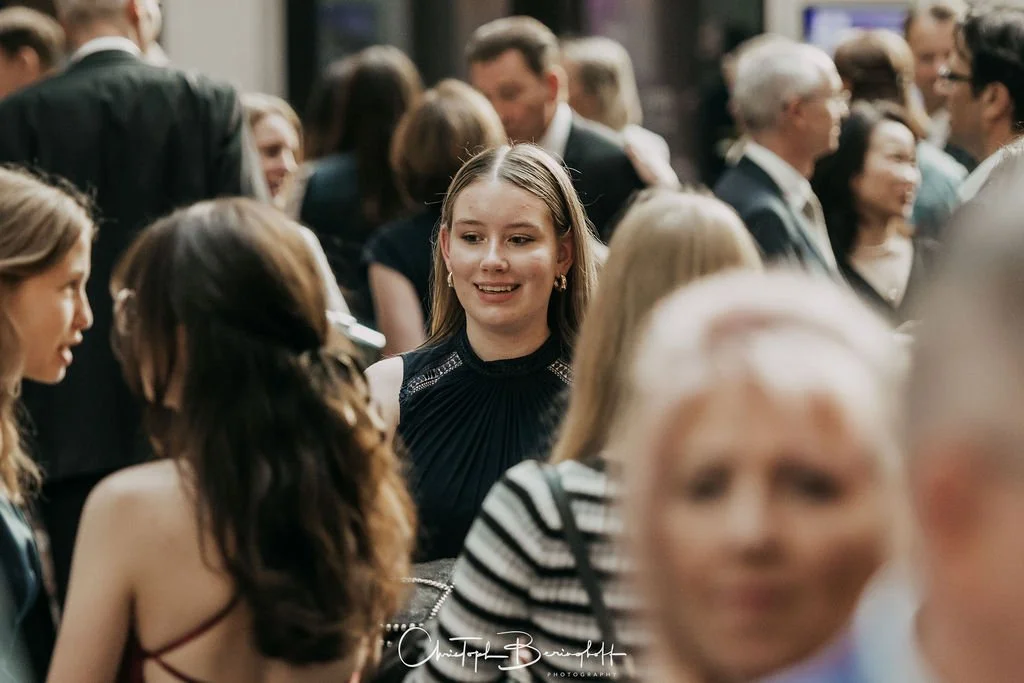 Menschen versammeln sich bei einer Veranstaltung und unterhalten sich. Im Mittelpunkt steht eine junge Frau mit blonden Haaren in einem schwarzen Kleid.