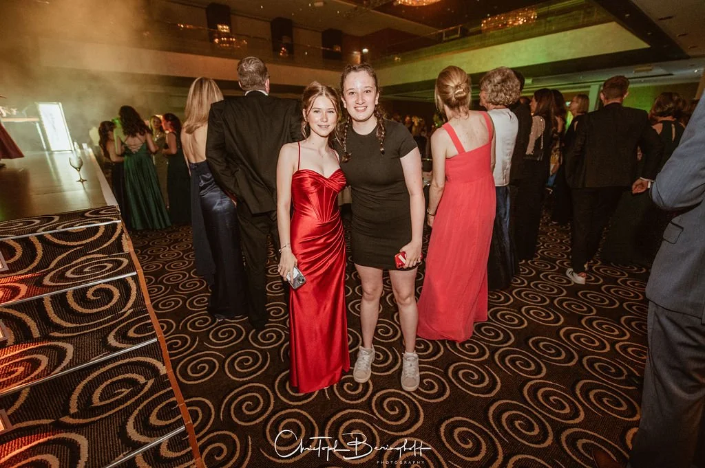 Zwei junge Frauen tanzen auf einer Party, eine trägt ein rotes Abendkleid, die andere ein schwarzes Kleid, umgeben von anderen festlich gekleideten Personen in einem großen festlichen Raum.