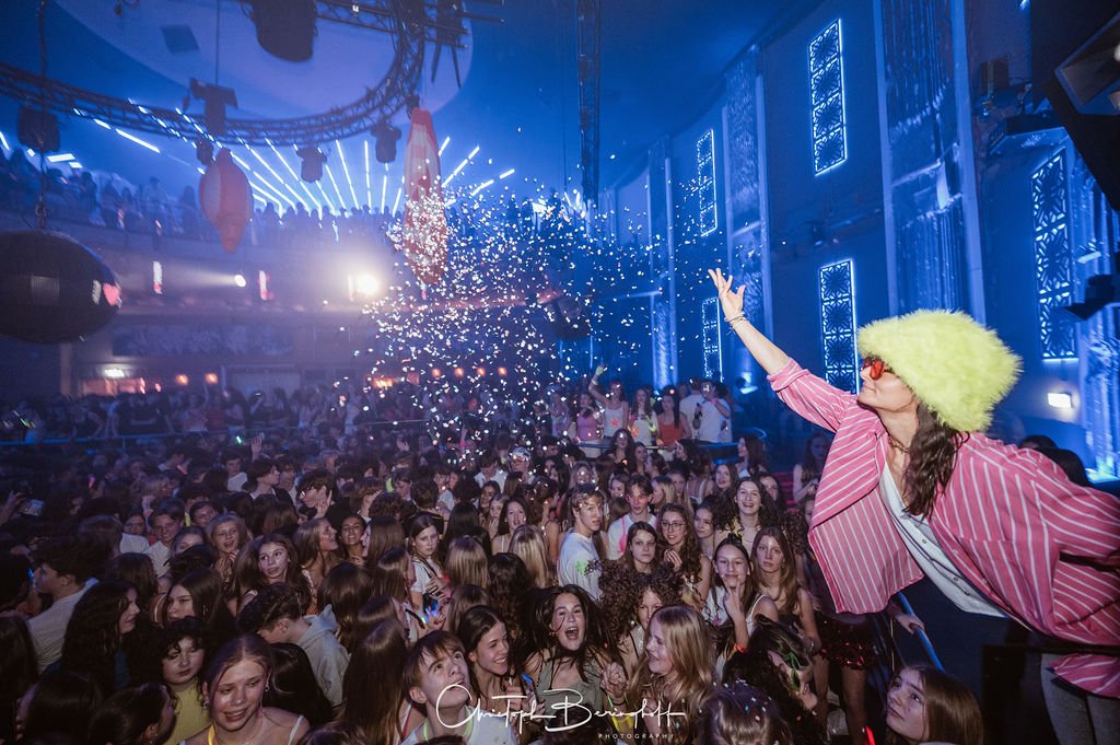 People are dancing at a party with colorful lighting effects; a woman wearing a yellow fluffy hat and sunglasses in the foreground is smiling and posing.