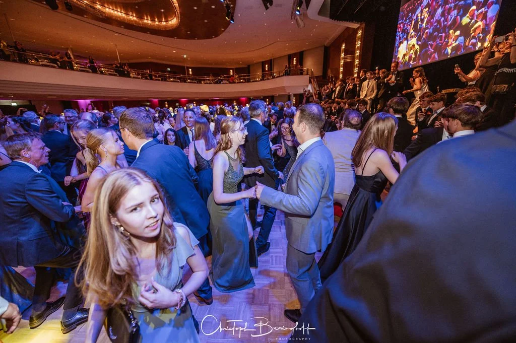 Menschen tanzen und feiern bei einer Gala oder Veranstaltung in einem eleganten Veranstaltungsraum mit großer Leinwand im Hintergrund.