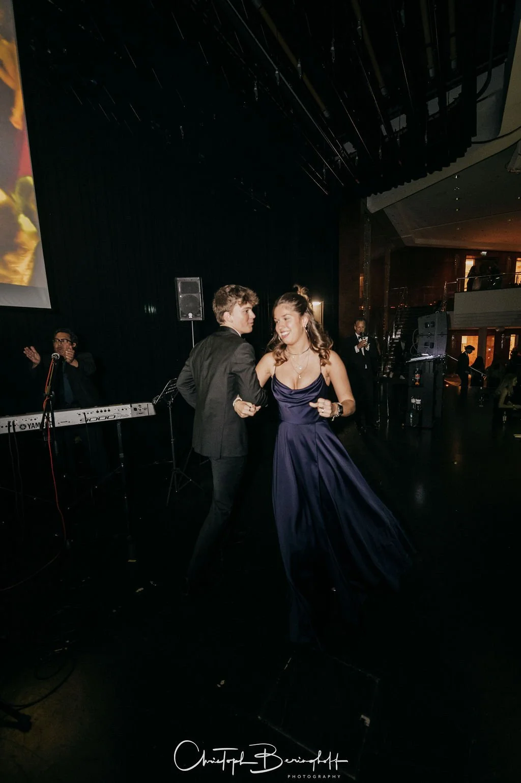 Ein junges Paar tanzt auf einer Party, die Frau trägt ein dunkles Abendkleid und der Mann einen schwarzen Anzug, im Hintergrund sind Musiker und dunkle Dekorationen sichtbar.
