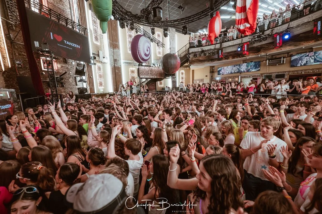 A large crowd at an indoor dance event or concert, with some large, colorful decorations hanging from the ceiling and a big screen displaying the text "I ❤️ DANCING."