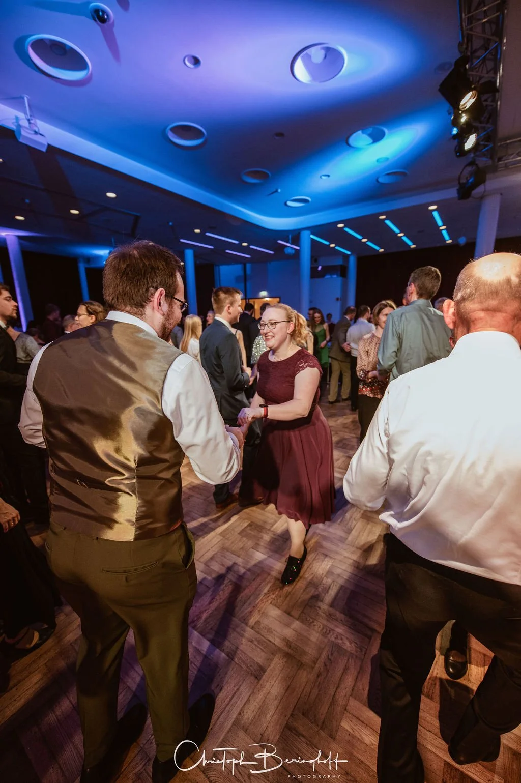 Menschen tanzen auf einer Party im Disco-Stil, mit blauer Beleuchtung und Holzparkett.