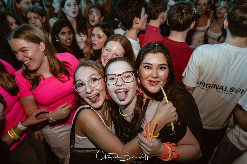 People laughing and having fun at a party, with a group of young women in the foreground sharing a hug.