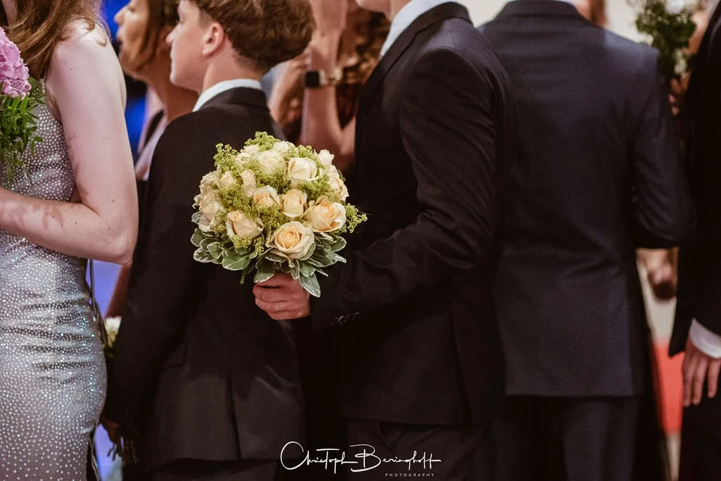 Junge in Schwarzanzug hält einen Blumenstrauß bei einer formellen Veranstaltung oder Hochzeit.