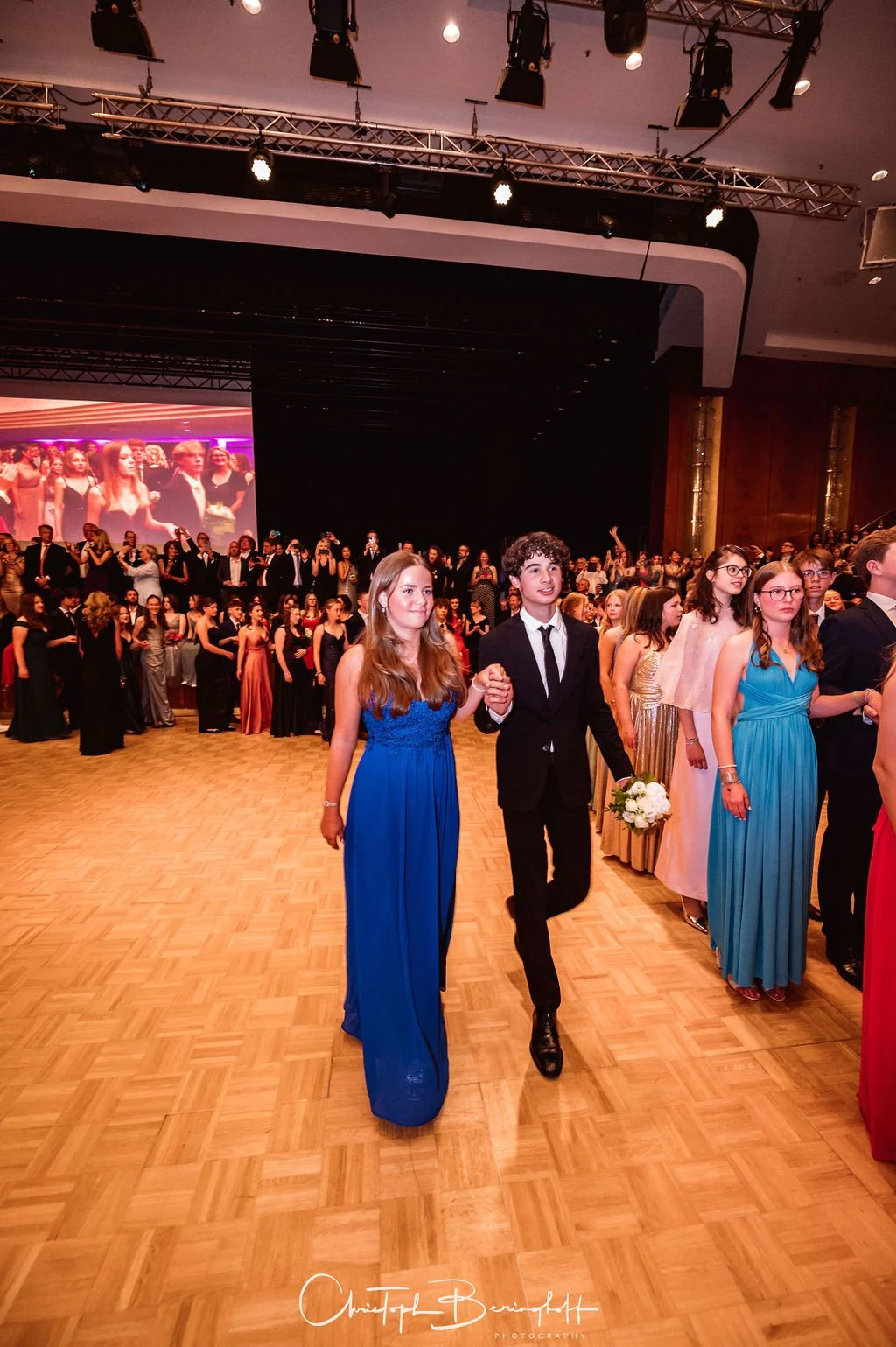 Junge Leute in eleganten Abendkleidern und Anzügen bei einer formellen Veranstaltung, vermutlich ein Ball oder Abschlussball, in einem großen Saal mit einer Tanzfläche und einer großen Projektion im Hintergrund.