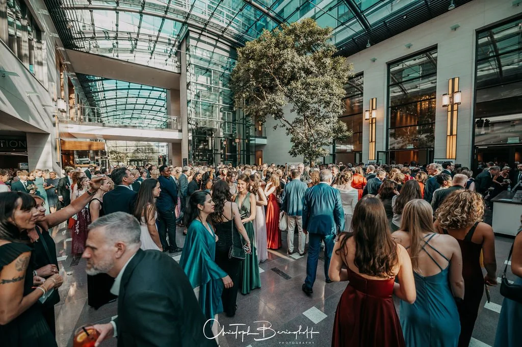 Menschen auf einer stilvollen Veranstaltung in einer modernen Halle mit Glasdach, viele in formeller Kleidung, manche sprechen, andere stehen in Gruppen, im Hintergrund ein großer Baum, helle Wanddekorationen.
