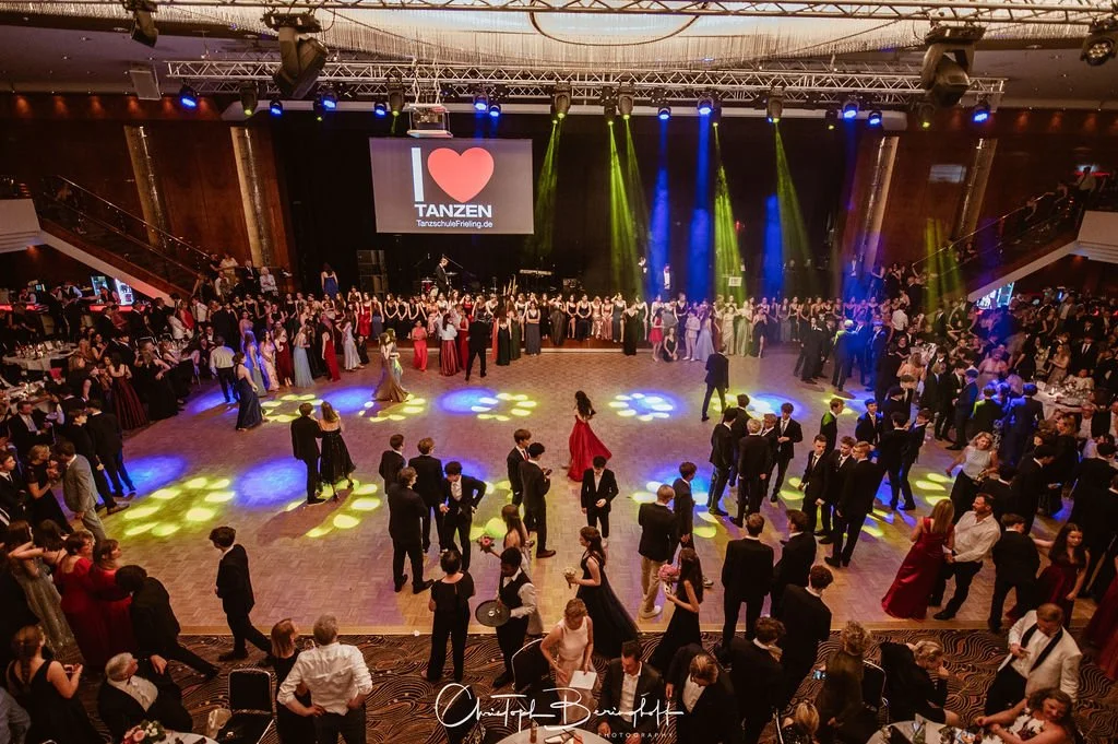 Tanzveranstaltung mit vielen tanzenden Paaren auf der Tanzfläche, im Hintergrund eine Bühne mit einer Leinwand, auf der steht "I ❤️ TANZEN", bunte Lichtstrahlen auf der Tanzfläche, Gäste in formeller Kleidung, im großen Saal mit Balustraden und Sitzg