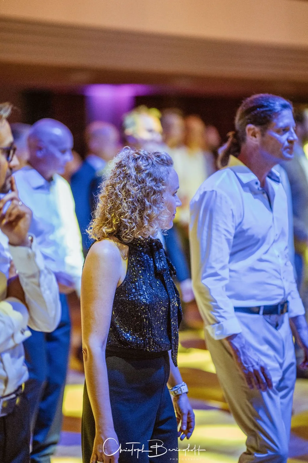 Menschen stehen in einer Gruppe bei einer Versammlung oder einem Event, bis auf eine Person sind alle mit weißen Hemden bekleidet, im Vordergrund eine Frau mit lockigem Haar und schwarzen Kleidung