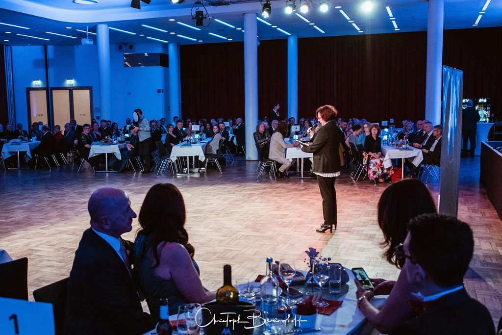 Menschen sitzen in einem festlich gedeckten Saal bei einem Event, einige hören einem Sprecher auf der Tanzfläche zu, auf der einige Personen sprechen.