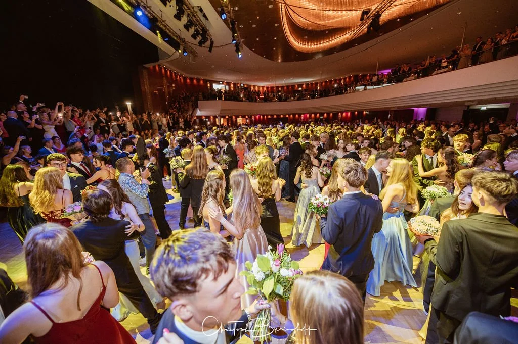Bilder von einem schwarzen-tie-Ball mit vielen jungen Menschen, die formelle Kleidung tragen, tanzen und sich mit Blumen in einem großen Saal mit Balkon und buntem Licht. Als Hintergrund sind Zuschauer im Obergeschoss zu sehen.