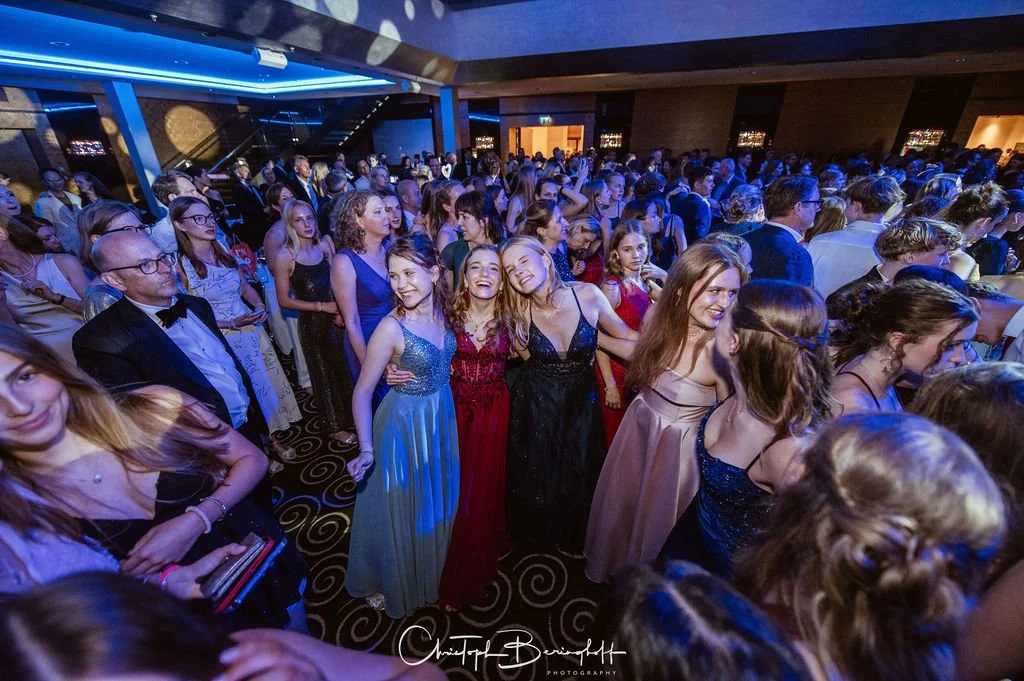 Viele junge Menschen in eleganten Kleidern tanzen auf einer Party in einem festlichen Raum mit blauer und warmer Beleuchtung.