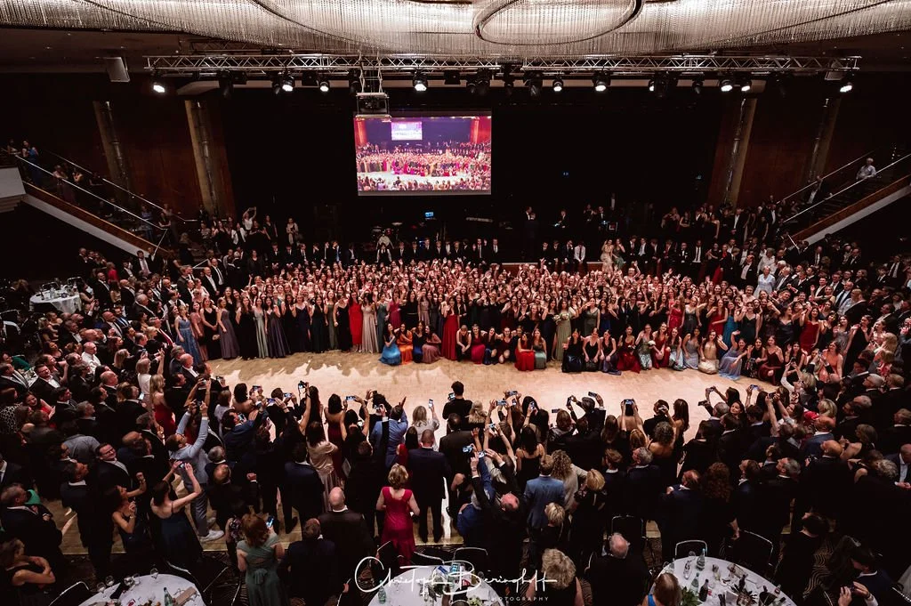 Großer Saal mit vielen Menschen bei einer formellen Veranstaltung, vermutlich einen Abschlussball oder eine Gala, mit Tanzfläche, Fotografen und einer großen Leinwand im Hintergrund.