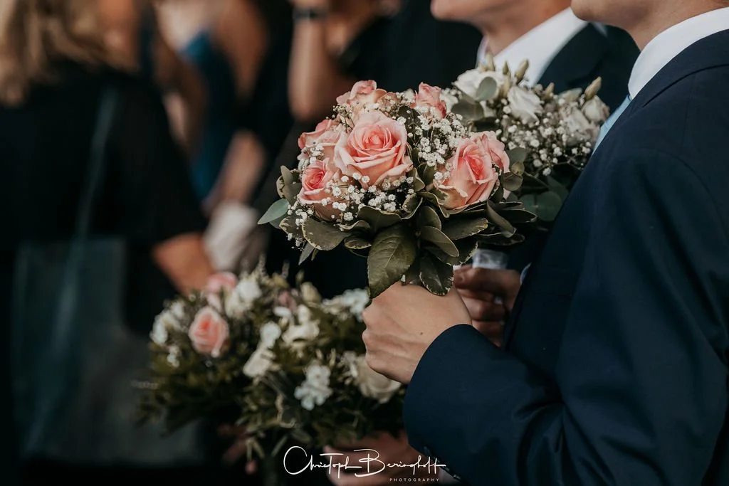 Ein Mann in Anzug hält einen Blumenstrauß mit rosa Rosen und weißen Blüten bei einer Hochzeit.