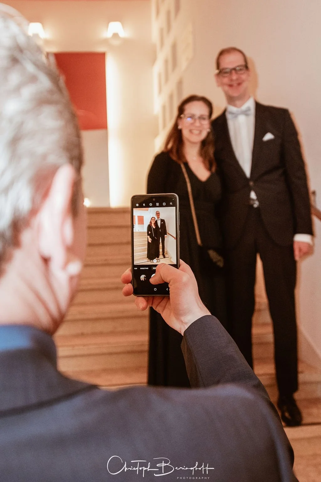 Ein Mann macht ein Foto von einem Paar, das in formeller Kleidung posiert, in einem hell erleuchteten Raum mit photographischer Wand im Hintergrund.