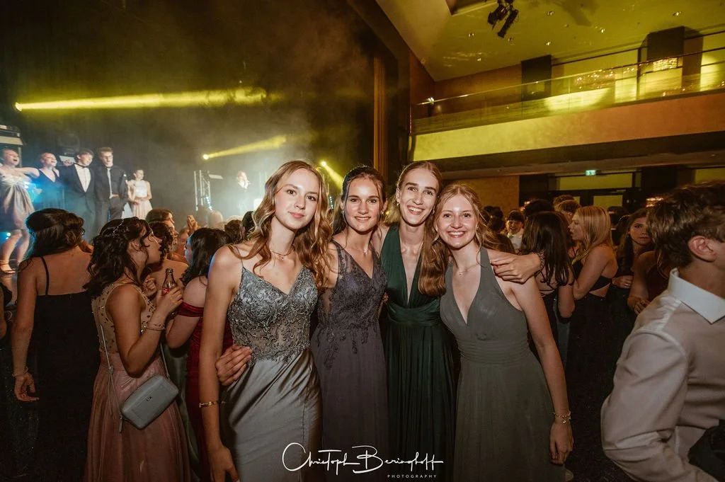 Vier junge Frauen in Abendkleidern posieren auf einer Party mit einer großen Menschenmenge im Hintergrund. Szenen aus einer festlichen Veranstaltung oder Ball.