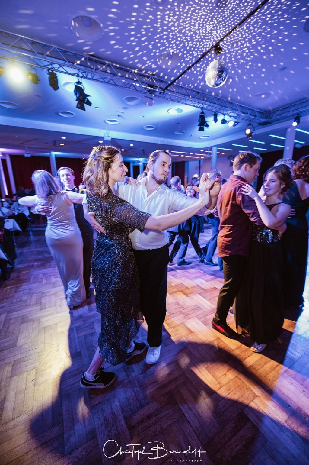 Paare tanzen in einem eleganten Raum mit Disco-Kugel und buntem Licht.