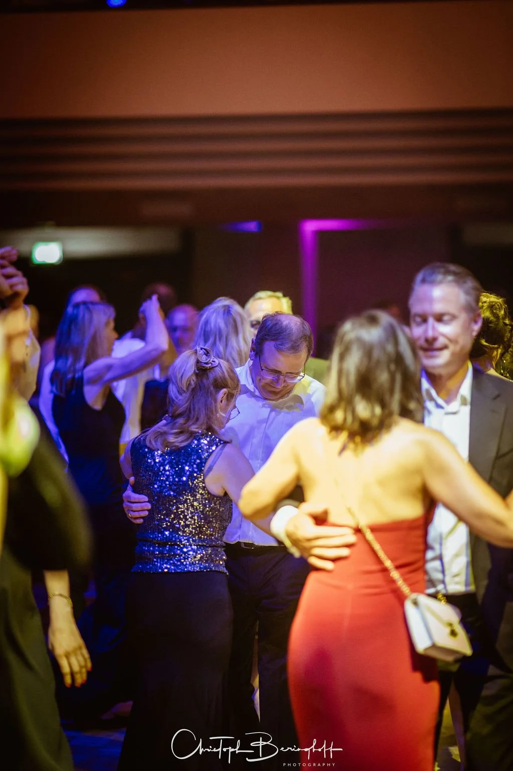 Menschen tanzen und feiern auf einer Party in einem dunklen Raum mit buntem Licht. Es gibt Männer und Frauen in formeller Kleidung, die umarmt und flirtend tanzen.