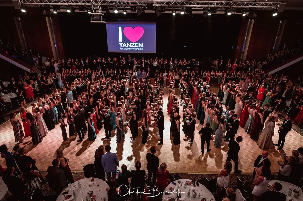 Tanzveranstaltung mit zahlreichen Paaren in formeller Kleidung tanzend auf einer großen, hell erleuchteten Tanzfläche. Im Hintergrund ein Bildschirm mit der Aufschrift "I ♥ TANZEN".