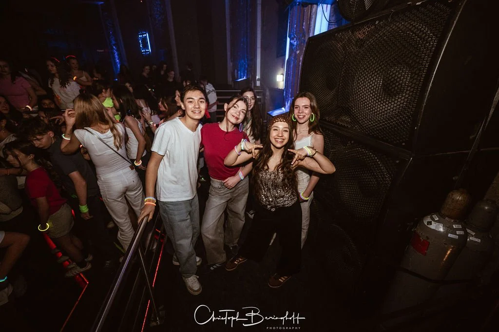 A group of young people in the middle of a club concert, posing in front of speakers, while other partygoers dance and celebrate in the background