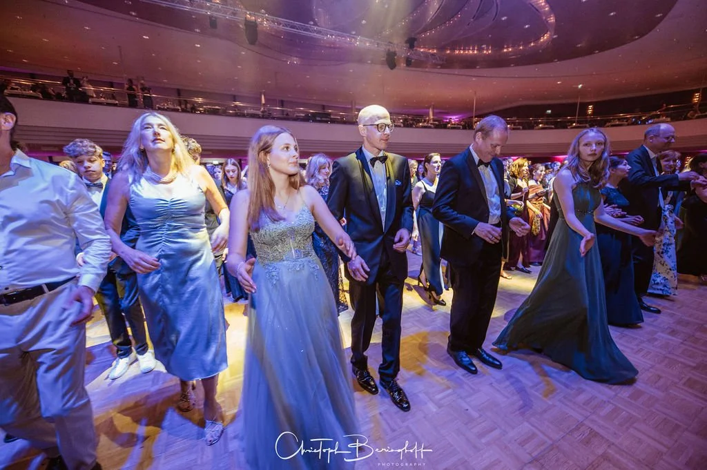 Menschen tanzen bei einer formellen Veranstaltung in einem großen Saal, die meisten in Gala-Outfits, mit einem gläsernen Deckenleuchter im Hintergrund.