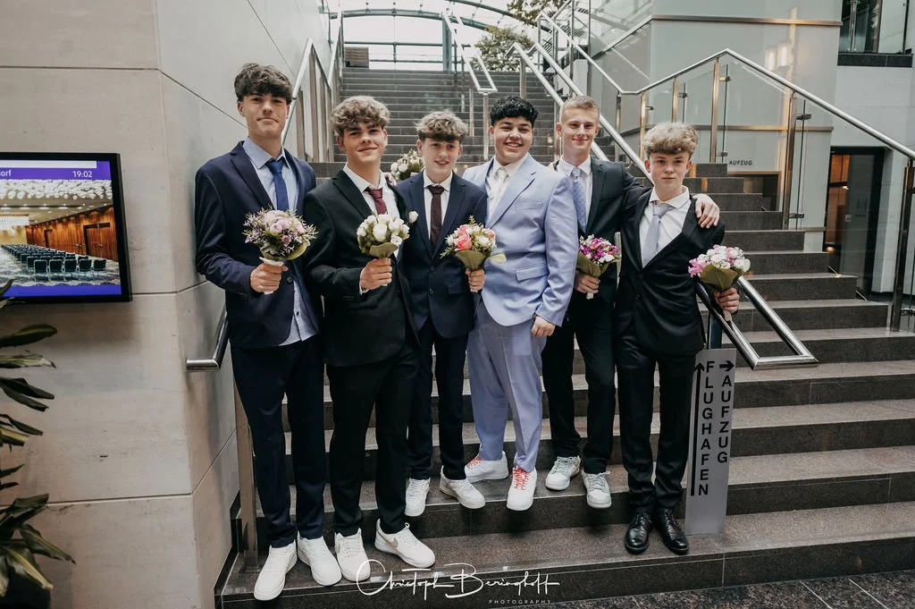 Sieben junge Männer in Anzügen stehen auf einer Treppe in einem modernen Gebäude, jeder hält einen Blumenstrauß. Es sieht nach einer offiziellen Veranstaltung oder Feier aus.