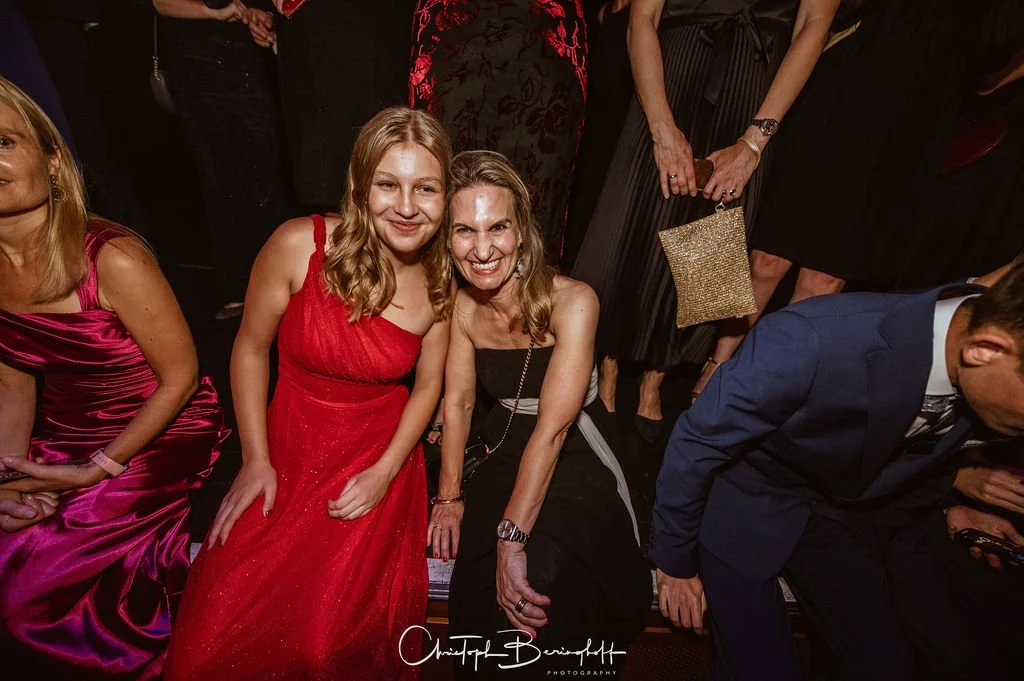 Zwei Frauen in eleganten Kleidern sitzen und posieren bei einer festlichen Veranstaltung. Viele Menschen sind im Hintergrund sichtbar, mit strahlenden Blicken und formeller Kleidung, in einer festlich dekorierten Umgebung.