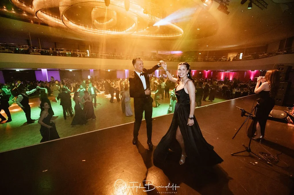 Paare tanzen auf einer Tanzfläche bei einer Feier, während ein Paar auf der Bühne tanzt, die Frau trägt ein schwarzes Abendkleid und der Mann einen Anzug, Licht- und LED-Effekte im Hintergrund.