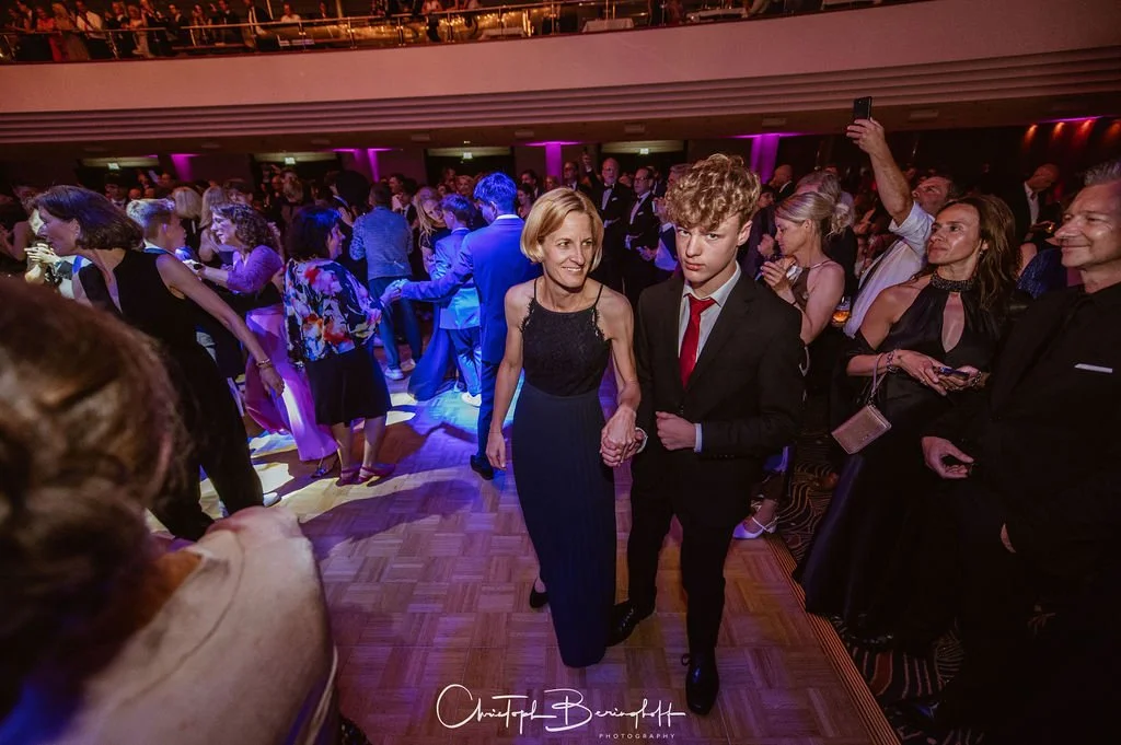 Menschen tanzen und feiern auf einem festlichen Event in einem eleganten Saal mit bunter Beleuchtung. Im Mittelpunkt eine ältere Frau und ein junger Mann in Anzügen, die sich die Hände halten, umarmt und auf der Tanzfläche.