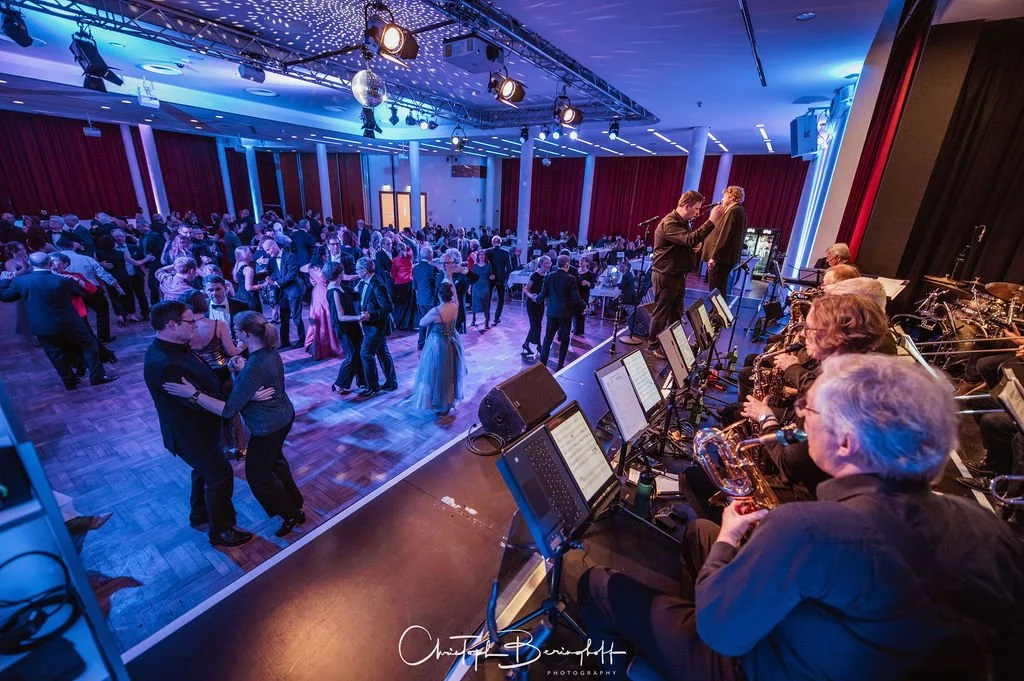Menschen tanzen auf einer Tanzfläche in einem festlich beleuchteten Saal, während eine Live-Band auf der Bühne spielt.