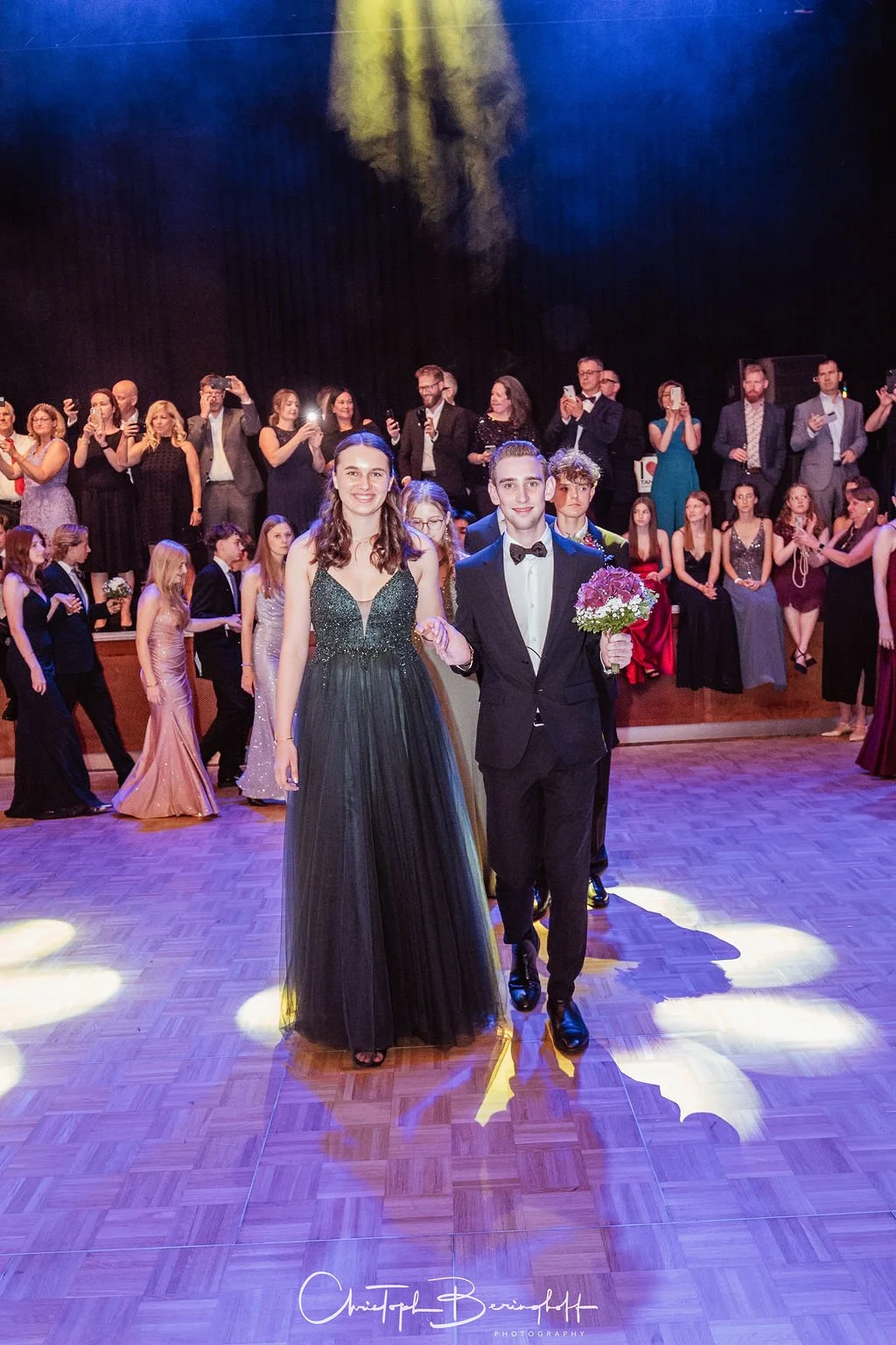 Ein junges Paar auf einem Abschlussball, die Frau trägt ein langes schwarzes Abendkleid, der Mann in einem schwarzen Anzug mit Fliege, der Mann hält einen Blumenstrauß, im Hintergrund stehen viele weitere Gäste und einige Fotografen.