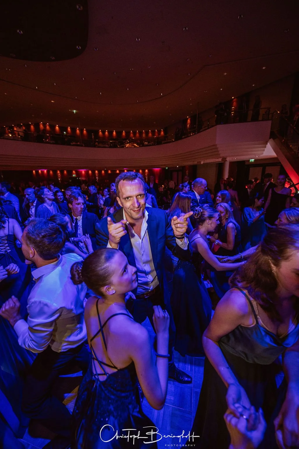 Menschen tanzen auf einer Party in einem großen Saal mit gedämpfter Beleuchtung, viele tragen elegante Kleidung.