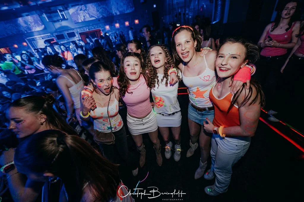 Groups of young people are dancing and partying in a dark room with blue lighting.