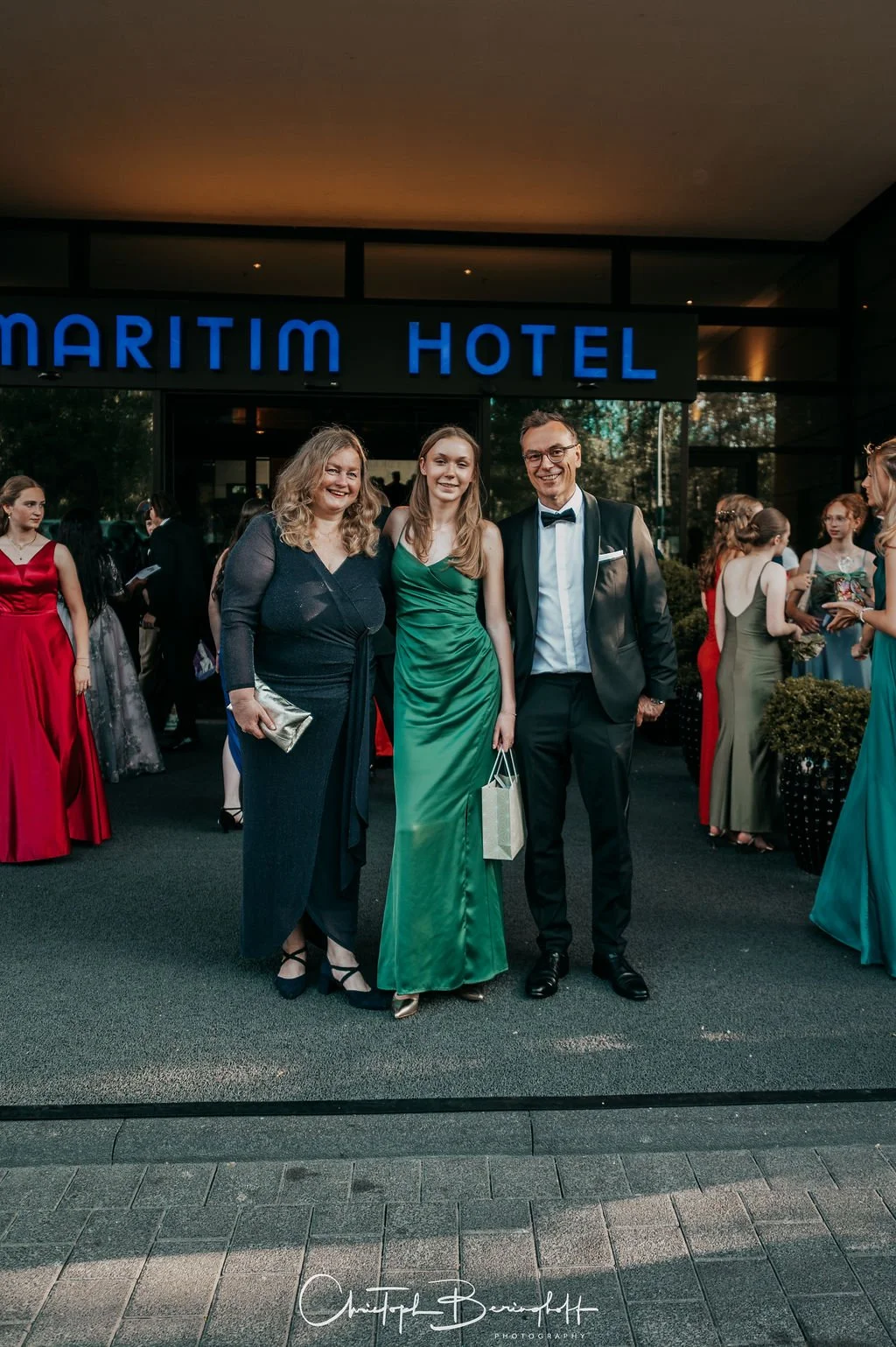 Drei Menschen in Abendgarderobe stehen vor dem Eingang des Maritim Hotels, umgeben von weiteren Gästen bei einer formellen Veranstaltung.