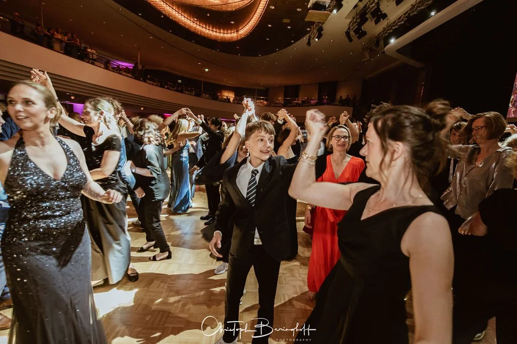 Menschen tanzen auf einer Feier im festlichen Abendkleid, in einem großen Saal mit heller Beleuchtung.