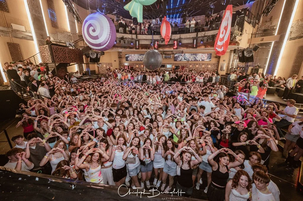 Many young people are dancing in a club and making heart shapes with their hands, including a large group in front of a dance floor, with a disco ball and colorful lights on the ceiling.