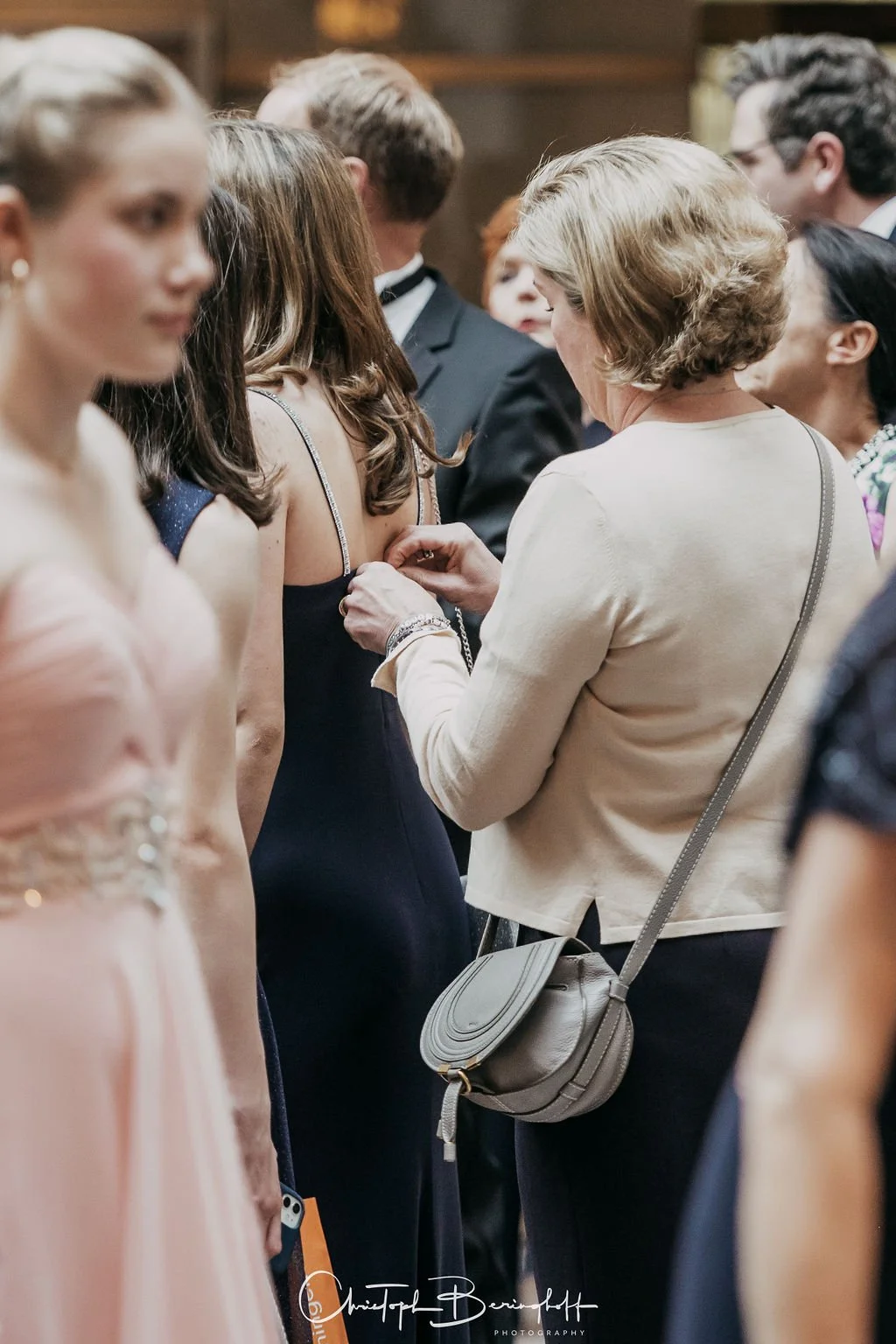 Eine Frau hilft einer jungen Frau, an einem formellen Anlass ein Schmuckstück anzulegen, umgeben von anderen gut gekleideten Menschen.
