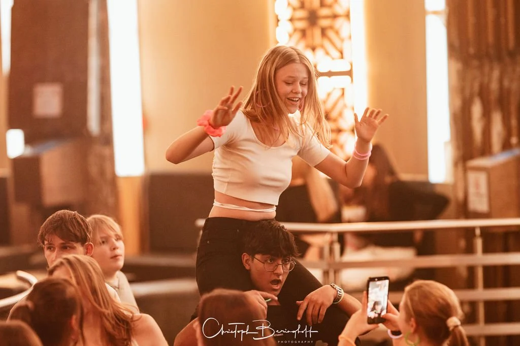 A young woman is sitting on a man's shoulders while she dances and laughs with other young people in a room.